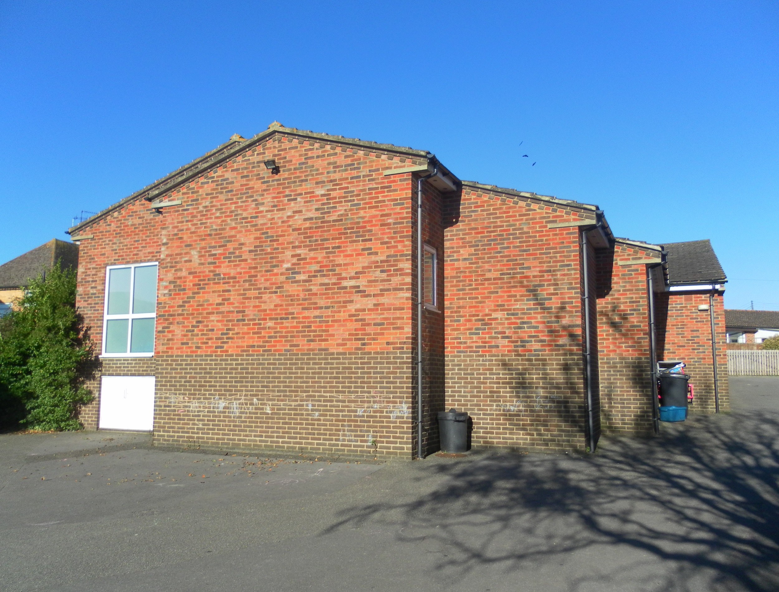 Rote Backstein-Semi-Detached-Bungalow mit weißer Tür und Fenstern, gelegen an der St. Johns Road, St. Johns Wood, Luton, umgeben von Bäumen und Mülltonnen, mit Himmel im Hintergrund.