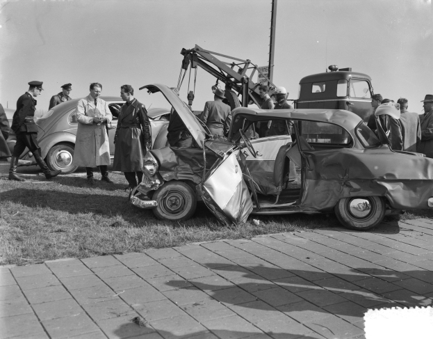Schwarzes und weißes Bild eines verunglückten Autos am Straßenrand mit mehreren Menschen drumherum und einem Mast im Hintergrund.