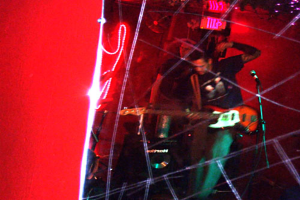 Ein Mann in schwarzem T-Shirt steht mit einer Gitarre in der Mitte, ein weiterer Mann hinter ihm spielt möglicherweise Instrumente, unter hellen Scheinwerfern, vor einer roten Wand bei einem Musikkonzert.