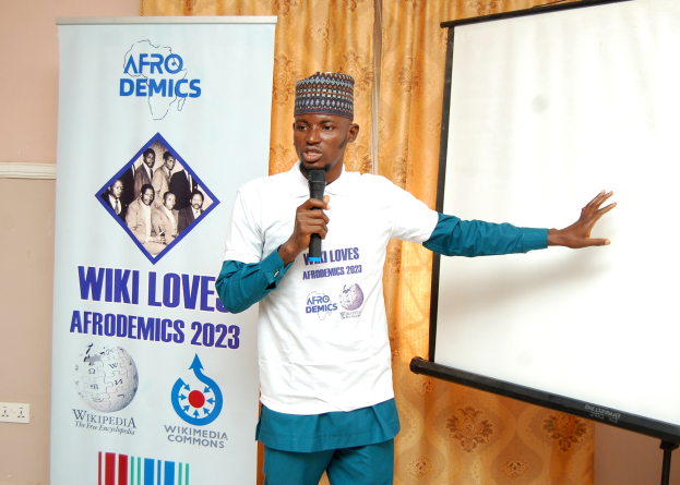 Ein Mann in einer Mütze hält ein Mikrofon und präsentiert vor einer Whiteboard, mit einem Banner, Vorhang und einer Schalttafel im Hintergrund.