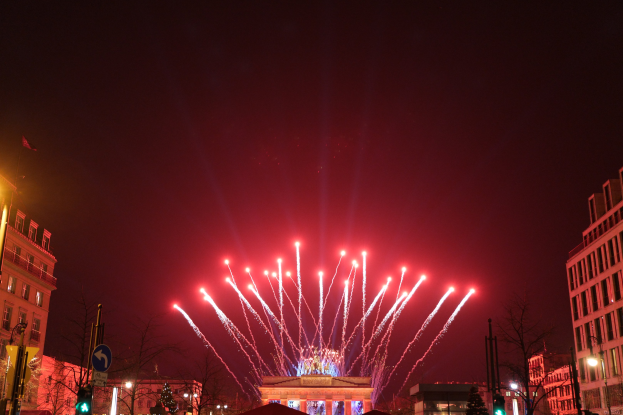 Das Bild zeigt eine nächtliche Stadtstraße in Berlin, Deutschland, mit dem Brandenburger Tor in der Mitte, umgeben von beleuchteten Gebäuden, Bäumen, Ampeln, Schildern und Fahnen, mit Feuerwerk, das den Himmel darüber erhellt.