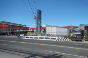 Tankstelle an einer Straßenkreuzung mit parkenden Autos, umgeben von Gebäuden, Versorgungsmasten, Schildern, Geländern und Pflanzen, unter einem bewölkten Himmel.