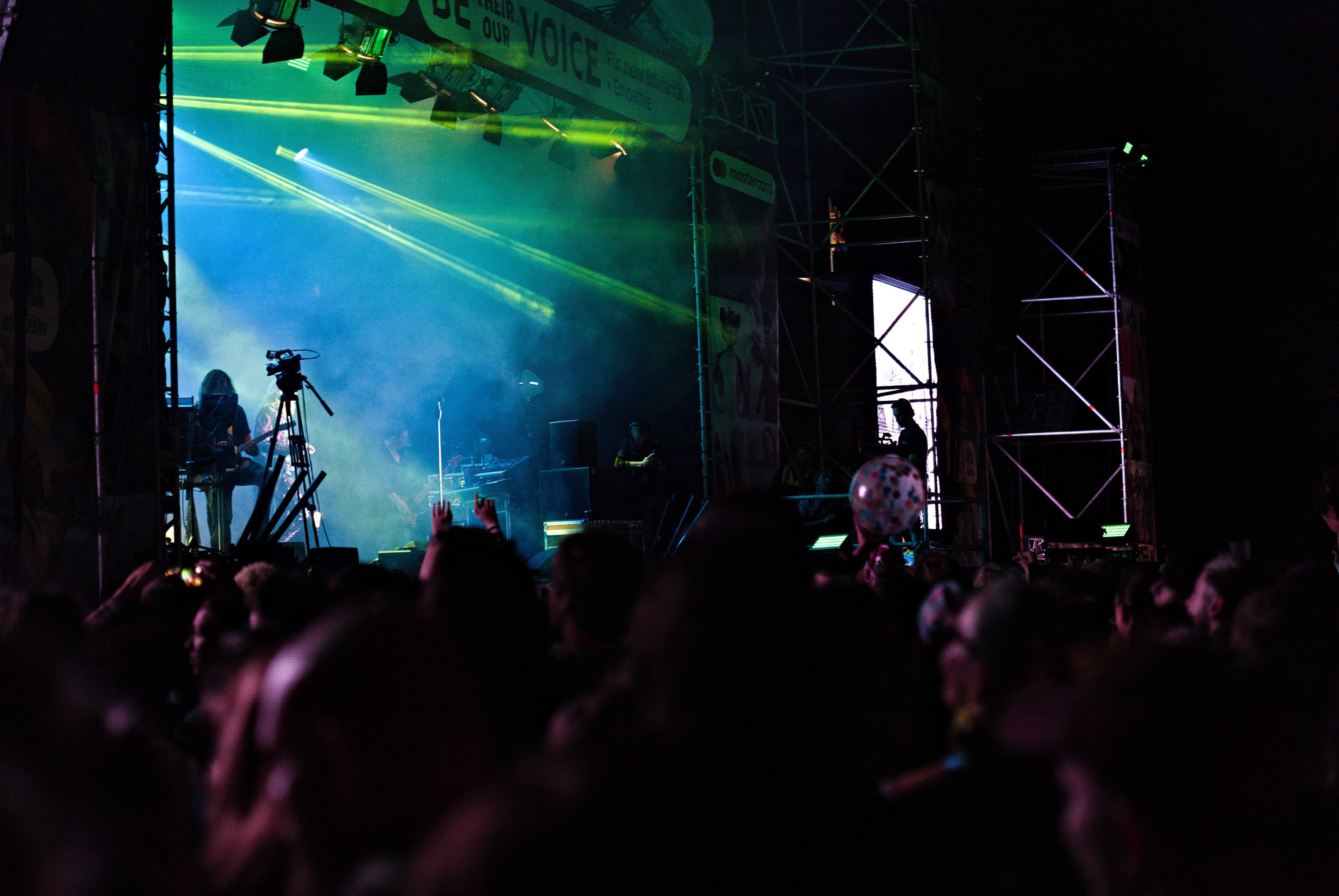 Ein volles Konzert mit Musikern auf der Bühne, beleuchtet von Scheinwerfern, mit Instrumenten, Mikrofonen und einem Banner im Hintergrund.