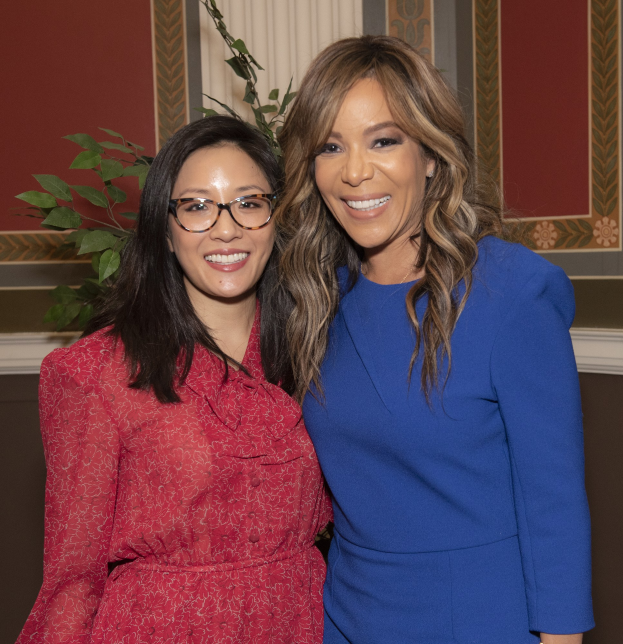 Zwei Frauen stehen nebeneinander und lächeln, eine trägt ein blaues Kleid und die andere ein rotes Kleid mit Brille, vor einer Wand mit einer Pflanze und einem Vorhang auf der 2019 Women in Film Pre-Oscar-Luncheon.