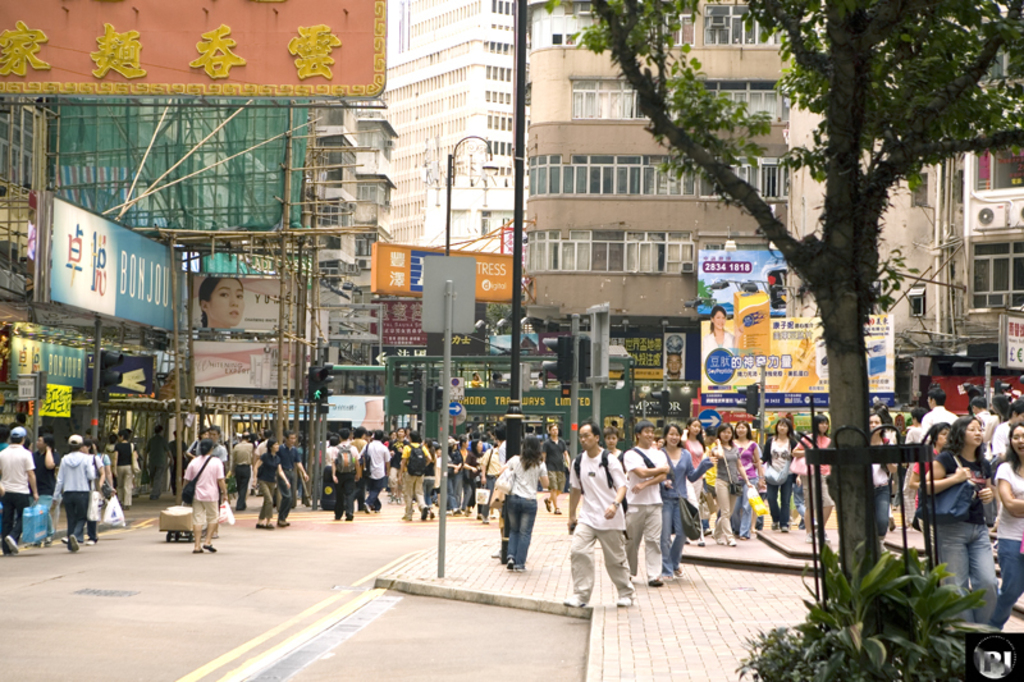 Eine belebte Straßenszene außerhalb der Stadt mit Menschen, Pfählen, Signalen, Plakaten, Pflanzen, einem Baum, Gebäuden, Rohren und einem entfernten Plakat im Hintergrund.