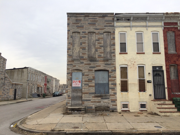 Ein altes, baufälliges Gebäude an einer Straßenecke mit zerbrochenen Fenstern, bröckelnden Wänden, vorbeifahrenden Fahrzeugen, Mülltonnen entlang des Bürgersteigs und einem klaren blauen Himmel.