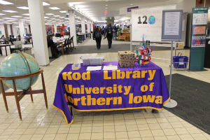 Eine Ausstellung mit einem Tisch, auf dem verschiedene Landesflaggen, eine Box, Papiere und ein Banner sowie links daneben eine Weltkugel, rechts eine Anschlagtafel mit einigen Menschen in der Nähe und mehrere Tische und Stühle weiter links zu sehen sind.