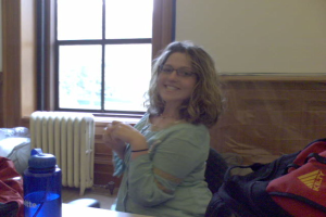 Eine Frau sitzt auf einem Stuhl drinnen mit einem Fenster im Hintergrund, einem Tisch mit einer Flasche, einer Jacke und einem Rucksack im Vordergrund und ein paar Dingen auf der linken Seite des Bildes.