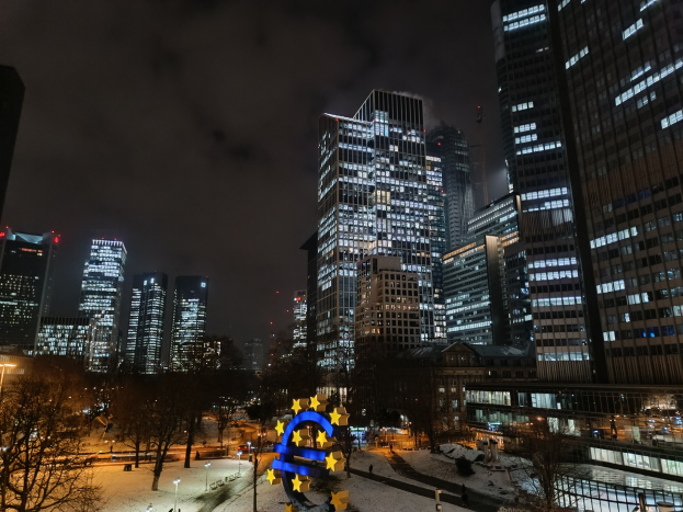 Hauptsitz der Europäischen Zentralbank in Frankfurt, Deutschland bei Nacht mit beleuchteten Gebäuden, Bäumen, Straßenlaternen, Lichtern und Fußgängern unter einem bewölkten Himmel.