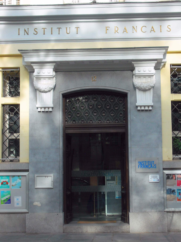 Eingang des Institut Français in Paris, mit einem Gebäude mit Fenstern, einer Tür, Plakaten an der Wand und einer Straße unten.