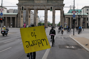 Eine Gruppe von Radfahrern mit Helmen fährt eine Straße vor dem Brandenburger Tor in Berlin, Deutschland, entlang, wobei einer ein gelbes Schild hält.