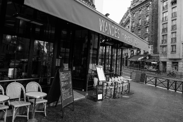 Ein Schwarz-Weiß-Bild einer Straßenszene mit einem Geschäft in der Mitte, Stühlen, einem Gebäude auf der rechten Seite, einer Straße und Fahrrädern.