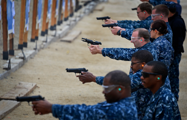 Eine Gruppe von Männern in Militäruniformen zielt mit Handfeuerwaffen auf Ziele auf der linken Seite des Bildes, mit einer Wand im Hintergrund.