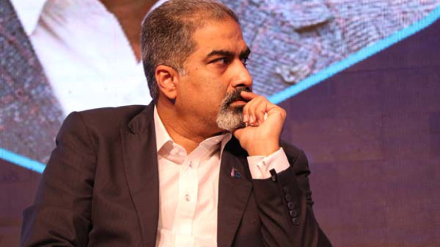 Ein Mann in Anzug und Krawatte sitzt auf einer Bühne, tief in Gedanken versunken, einem Bildschirm zugewandt.