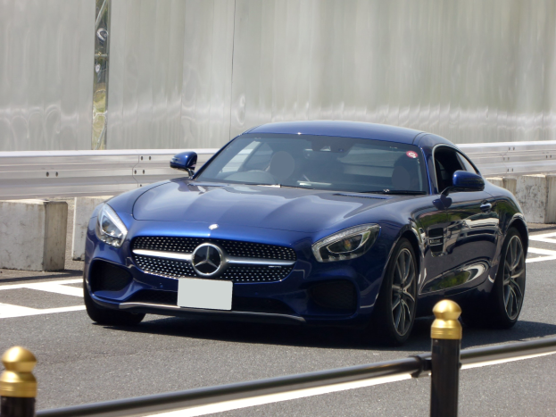 Blauer Mercedes-Benz AMG GT Coupé auf einer Straße unterwegs, mit einem schlanken aerodynamischen Design mit eckigen Scheinwerfern, einem großen Kühlergrill, glänzenden großen Rädern und einem luxuriösen Lederinnenraum mit einem modernen Armaturenbrett, vor einer Wand.