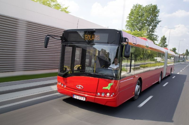 Ein roter Bus fährt auf einer Straße neben einem Gebäude, mit einer Person im Inneren, neben einer Wand, mit Bäumen, Laternen und einem bewölkten Himmel im Hintergrund.