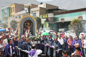 Menschen auf einer Straße außerhalb der Stadt, mit einem Kreuz und Blumensträußen im Vordergrund, Autos im Hintergrund und Gebäuden, Bäumen und Straßenlaternen weiter hinten.
