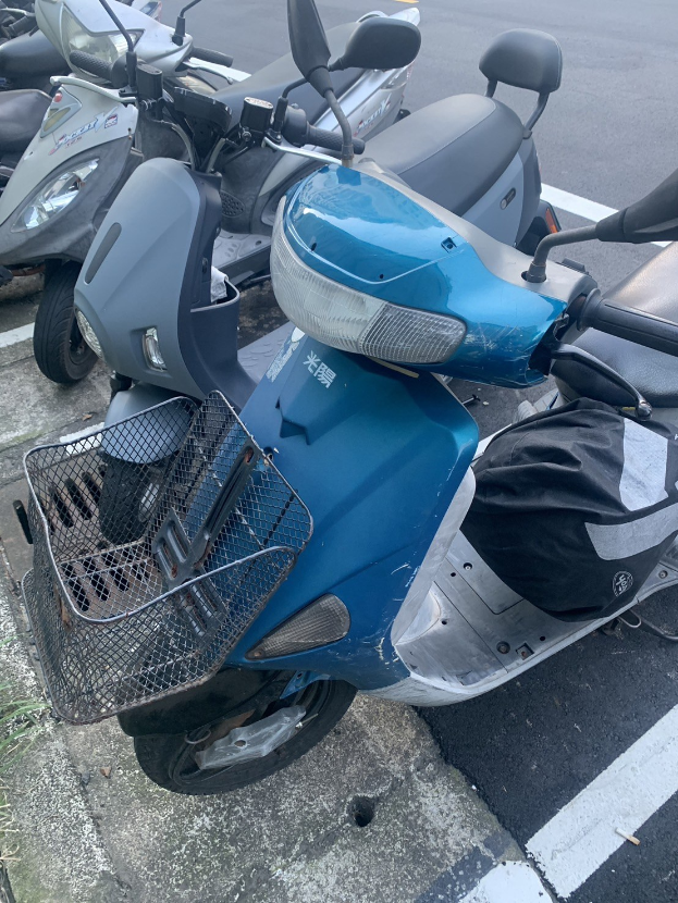 Ein blaues Roller auf einer Straße neben anderen Motorrädern geparkt, mit Gras links sichtbar.