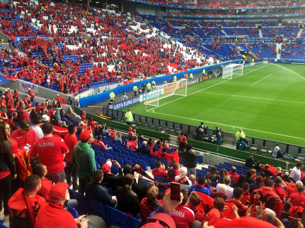 Eine große Menschenmenge in einem Stadion schaut einem Fußballspiel zu, mit Menschen auf Sitzen und Stehplätzen, Bannern, einem Torpfosten und Deckenleuchten unter einer Dachkonstruktion.