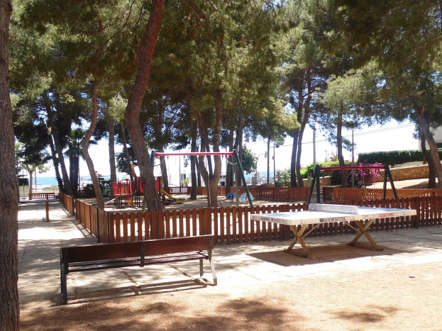 Eine Parkszene mit einem zentralen Tischtennis-Tisch, Bänken, Schaukeln, Bäumen und einem Zaun, mit einem Wasserlauf und einem blauen Himmel im Hintergrund.