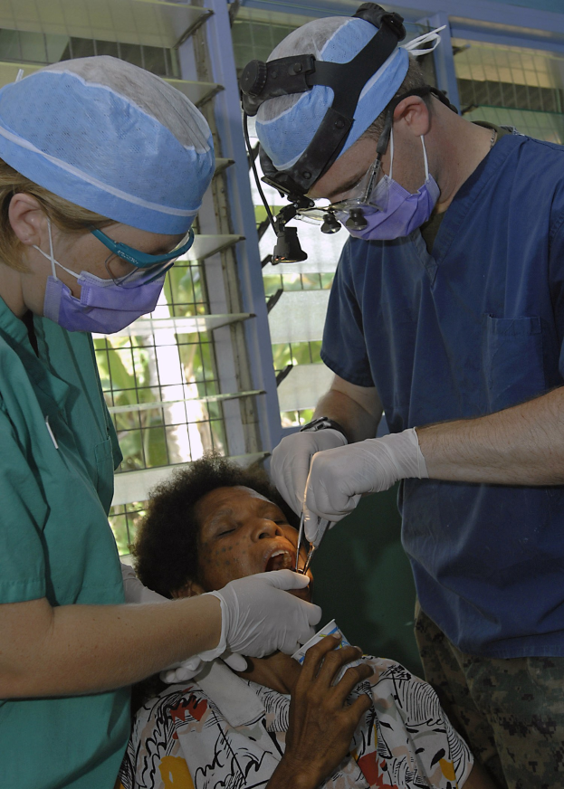 Zwei Chirurgen in Masken und Kappen führen eine Gesichtsoperation an einer Frau durch, die auf einem Bett liegt, während einer ein chirurgisches Instrument hält und ein Fenster mit Bäumen im Hintergrund zu sehen ist.