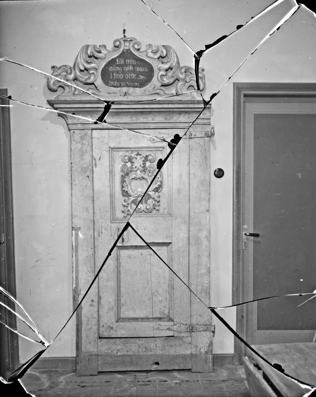 Ein Schwarz-Weiß-Foto einer zerbrochenen Tür in einem Raum mit Glasscherben drumherum und einer Wand im Hintergrund.