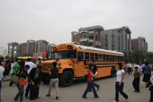 Eine Gruppe von Menschen, einige mit Taschen, geht vor einem Schulbus auf einer Straße, die von Gebäuden, Bäumen und Laternen gesäumt ist, unter einem klaren blauen Himmel.
