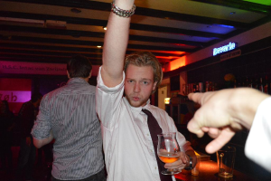 Eine Gruppe von Menschen, die in einer Bar stehen, mit einem Mann in einem weißen Hemd und schwarzer Krawatte, der ein Weinglas in der Nähe der Bartheke hält, auf der viele Weinflaschen und "Bayern" darüber geschrieben stehen.