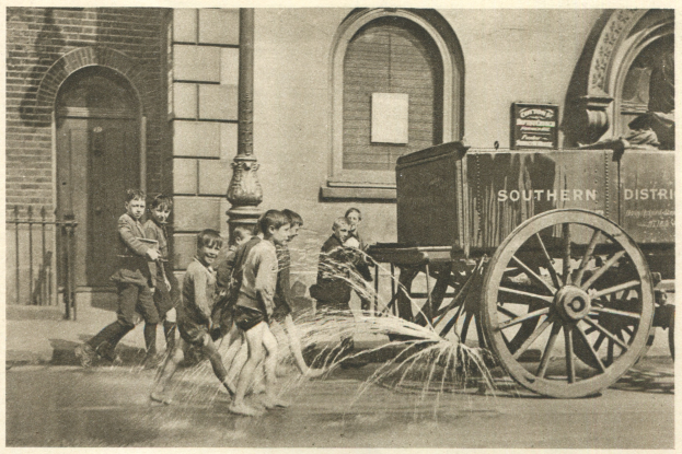 Schwarz-weißes Foto von Kindern, die mit einem Feuerhydranten vor einem Gebäude mit Fenstern, einer Tür, einem Mast und einem Zaun spielen, mit einem Wagen im Hintergrund.