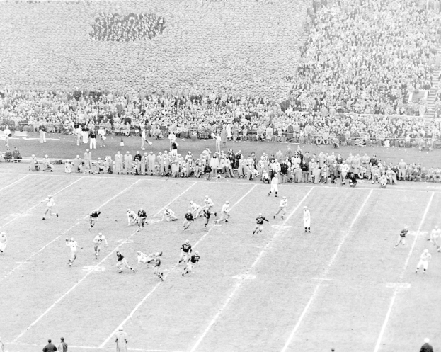 Schwarzes und weißes Foto eines Fußballspiels im Gange, mit Spielern auf dem Boden und Zuschauern im Hintergrund.