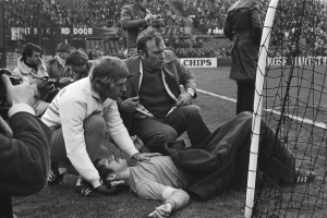 Eine Schwarz-Weiß-Szene eines Mannes, der neben einem Fußballtor auf dem Boden liegt und von einer Gruppe von Menschen auf dem Rasen umgeben ist, mit einem Netz auf der rechten Seite und Zuschauern in den Rängen, einige halten Kameras, und Tafeln mit Text im Hintergrund.