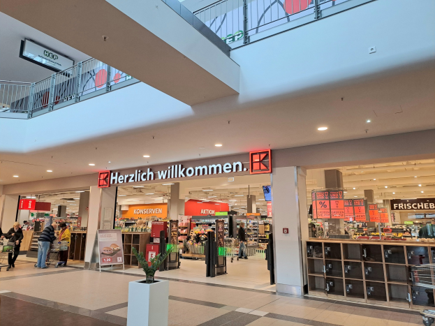 Innenansicht eines Einkaufszentrums mit Menschen, Geschäften, Informationsschildern, Beleuchtung, Geländern und einem Topf mit einer Pflanze am Boden; ein Schild trägt die Aufschrift "Herzlich willkommen", was auf einen deutschen Supermarkt hinweist.