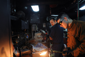 Zwei Männer mit Brille arbeiten an einem Metallstück in einer Werkstatt, einer verwendet ein Schweißgerät, mit verschiedenen Werkzeugen und Equipment auf dem Tisch und einem Fernseher und Lichtern im Hintergrund.