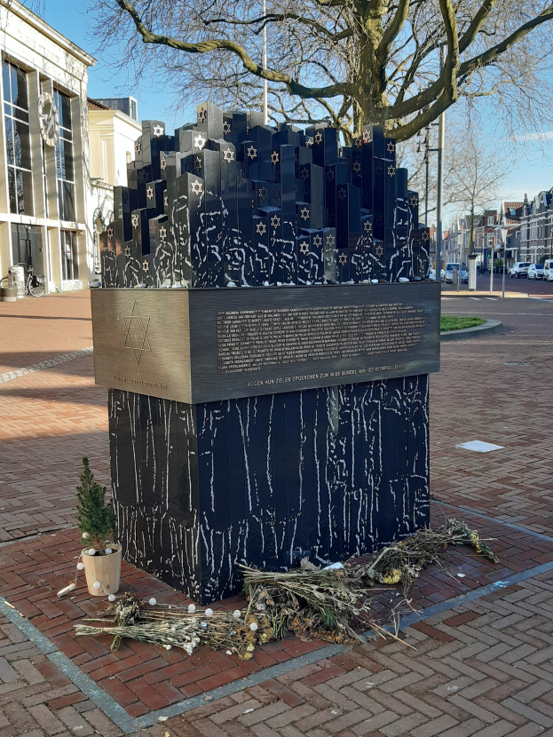 Denkmal in einem Stadtplatz, das an Holocaust-Opfer erinnert, mit einem Baum davor, einer Topfpflanze auf dem Boden, verstreuten trockenen Blättern und im Hintergrund Gebäude, Fahrzeuge, Masten und Bäume.