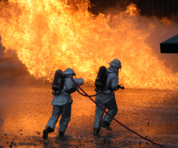 Zwei Feuerwehrleute in Schutzausrüstung mit einer Schlauchleitung ein Feuer löschend, mit einem Objekt rechts im Bild.