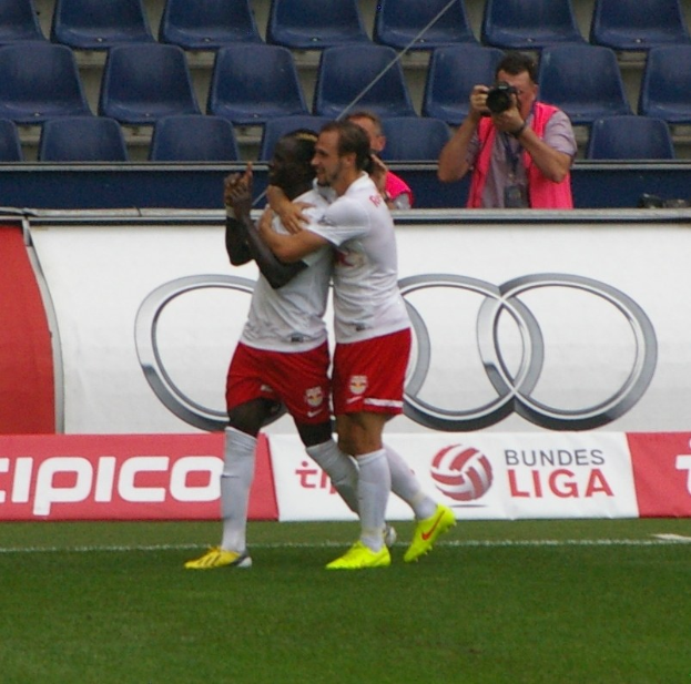 Zwei Fußballspieler feiern ein Tor auf einem Feld mit einer Menge, einem Fotografen, einer Fahne, Stühlen und einer Wand im Hintergrund.