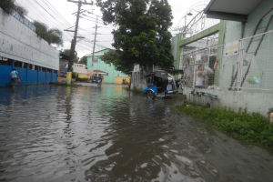 Eine überflutete Straße mit einem geparkten Motorrad in der Mitte, flankiert von Gebäuden, Bäumen und Strommasten mit Drähten und einigen Menschen und Fahrzeugen im Hintergrund.