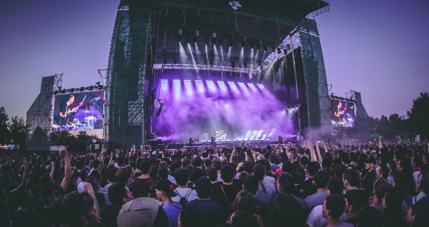 Eine große Menschenmenge schaut bei einem Musikfestival auf einer beleuchteten Bühne mit Instrumenten und Bildschirmen, die vor einem Gebäude, Bäumen und einem klaren blauen Himmel steht.