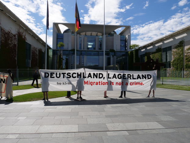 Gruppe von Menschen mit einem Banner, auf dem "Deutschland, Lagerland Migration ist kein Verbrechen" steht, vor einem Gebäude mit Fenstern, umgeben von Gras, einem Metallzaun, Pflanzen, Bäumen und Fahnen auf Stangen unter einem bewölkten Himmel.