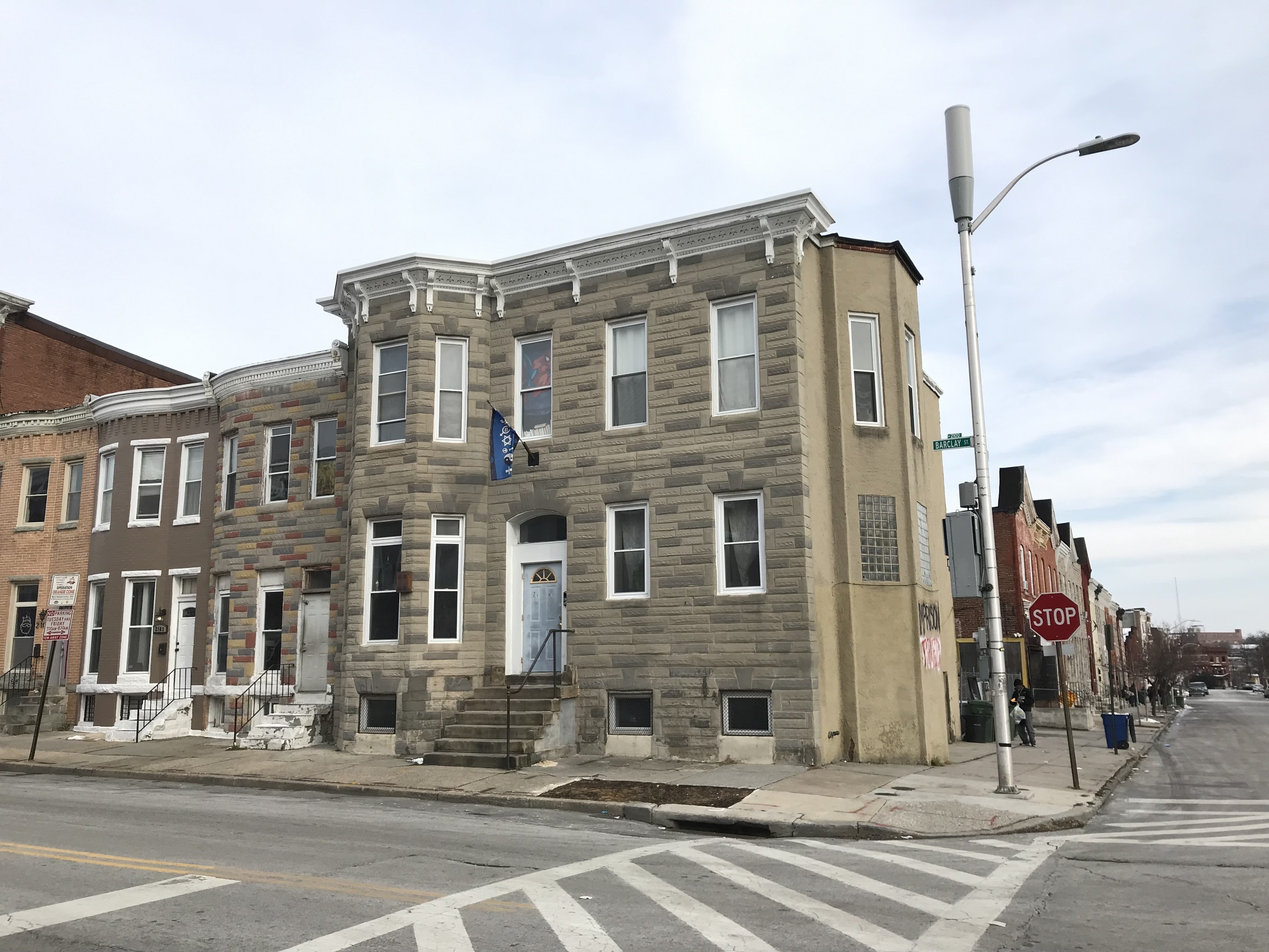Eine Häuserreihe an einer Straßenkreuzung mit Fahrzeugen, Laternen, Schildern, Bäumen und einem bewölkten Himmel im Hintergrund.