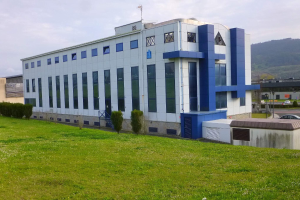 Großes weißes und blaues Gebäude mit Fahrzeugen, Masten und Bergen im Hintergrund, auf einem grünen Feld mit Bäumen und Gras unter einem klaren blauen Himmel.