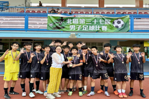 Eine Gruppe junger Männer in Fußballtrikots steht zusammen auf einem Feld, hält einen Pokal und trägt Medaillen, mit einem Banner im Hintergrund, das 'Yokohama U-16 Jungenfußballteam' lautet, und ein paar Menschen sitzen vor einem Gebäude.