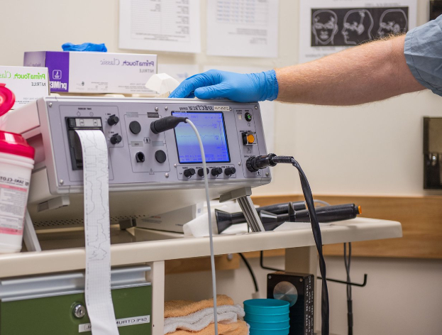 Eine Person in blauen Handschuhen arbeitet an einem medizinischen Gerät auf einem vollen Tisch in einer Krankenhausumgebung mit Papieren an der Wand im Hintergrund.
