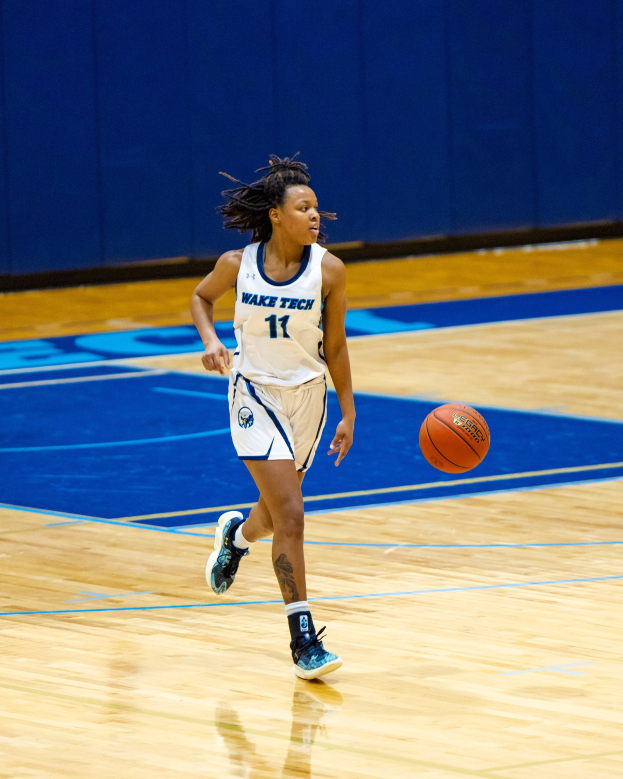 Eine Frau in einer blauen und weißen Uniform, die einen Basketball auf einem Court dribbelt, trägt ein weißes T-Shirt mit der Aufschrift "Wake Tech Women's Basketball" und blaue Schuhe, mit einer blauen Wand im Hintergrund.