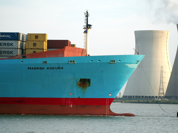 Großes blaues und rotes Containerschiff mit gestapelten Containern auf dem Wasser in der Nähe eines Atomkraftwerks mit sichtbarer Kühlturm im Hintergrund unter einem klaren Himmel.