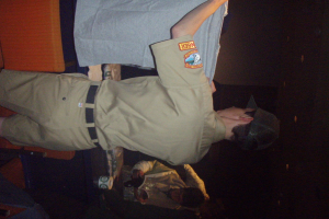 Ein Polizeibeamter hält ein T-Shirt in den Vordergrund, während im Hintergrund ein Junge ein Buch und einen Becher hält und in der Nähe eines Schreibtischs und Lampen steht.