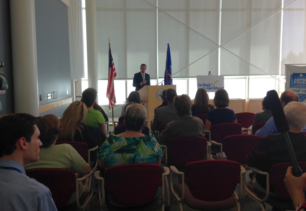 Ein Mann steht an einem Podium und spricht zu einem Publikum, das auf Stühlen sitzt, mit zwei Fahnen daneben und einer Wand, Säulen und Fenstern im Hintergrund.