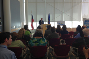 Ein Mann steht an einem Podium und spricht zu einem Publikum, das auf Stühlen sitzt, mit zwei Fahnen daneben und einer Wand, Säulen und Fenstern im Hintergrund.