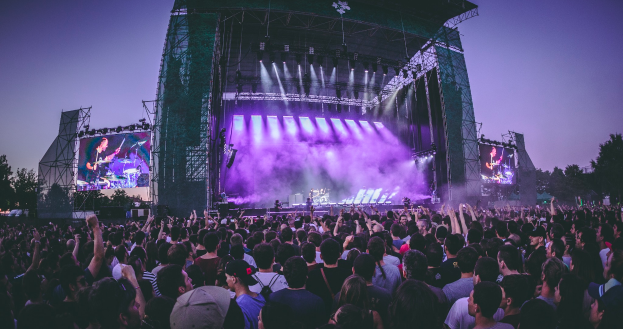 Eine große Menschenmenge auf einem Musikfestival, mit einer Bühne in der Mitte, auf der Musiker Instrumente spielen, beleuchtet von Scheinwerfern und umgeben von Bildschirmen, mit einem Gebäude, Bäumen und einem klaren blauen Himmel im Hintergrund.