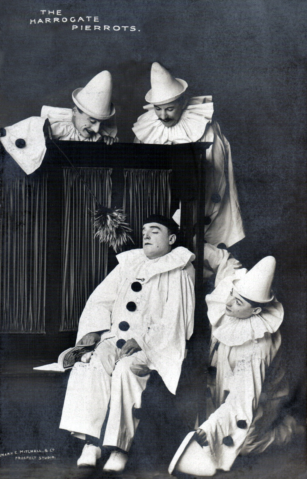 Ein Schwarz-Weiß-Foto einer Gruppe von Clowns, die auf einer Bühne sitzen, wobei eine Person ein Buch hält und Vorhänge im Hintergrund zu sehen sind.